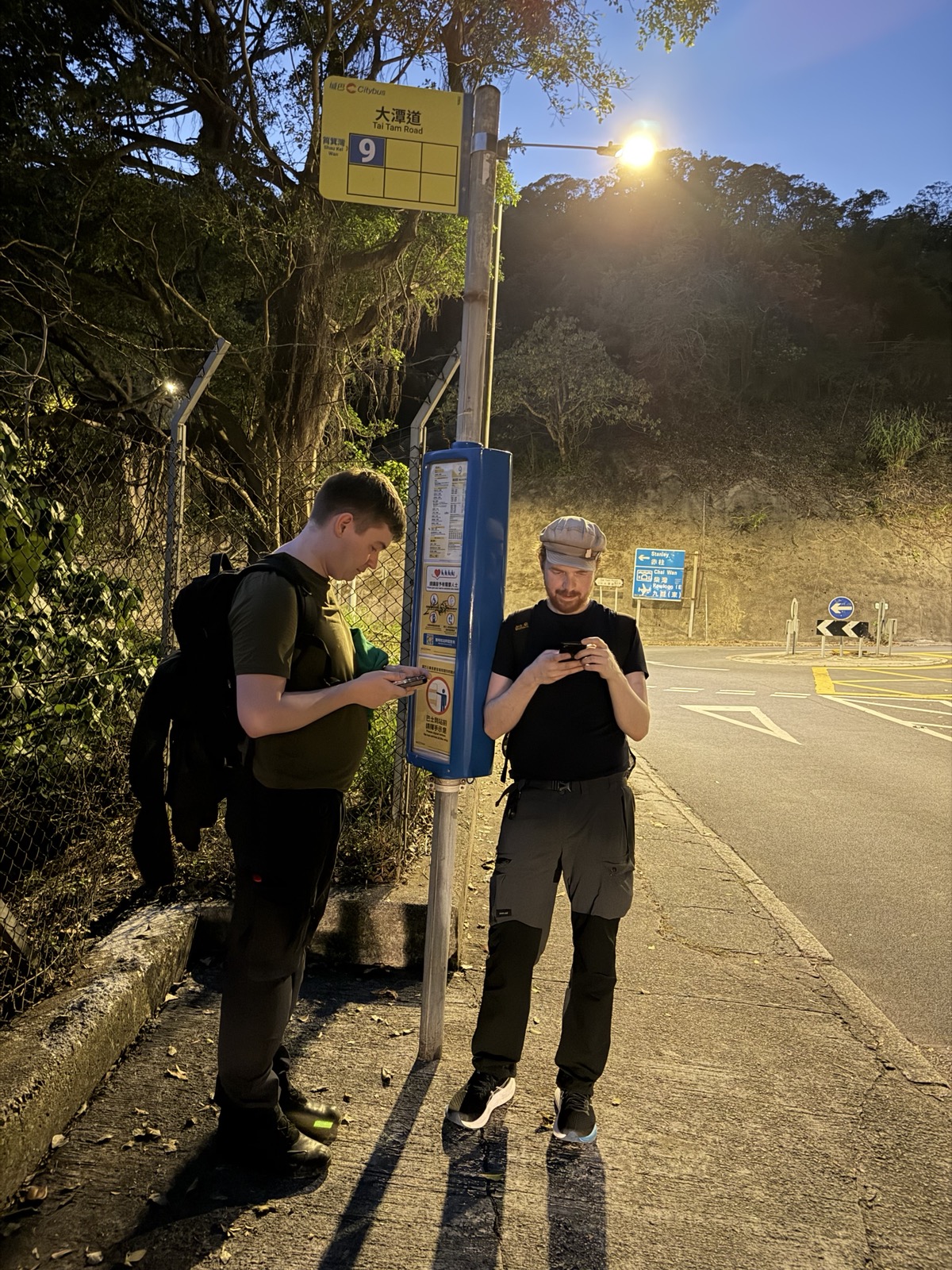 Two people stand under a bus stop sign, looking at their smartphones.
