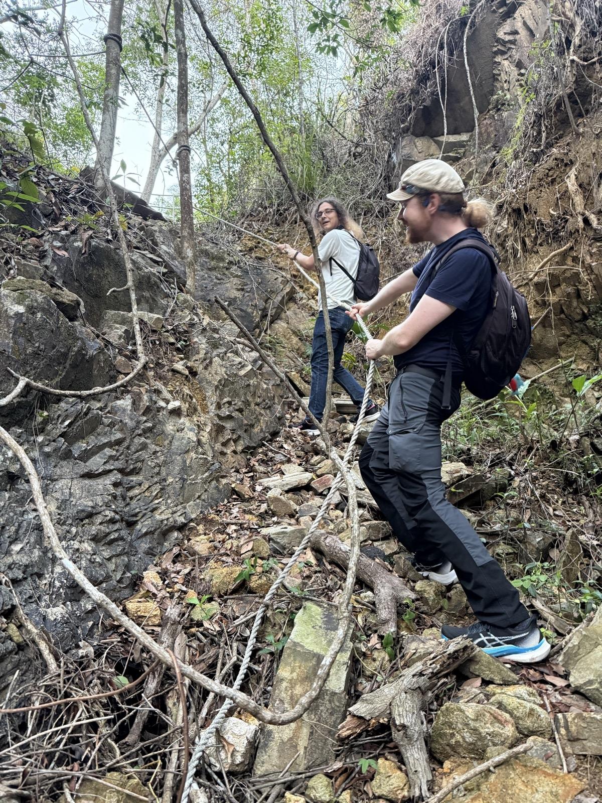 Two people pull themselves up a steep stretch of path using a rope that's knotted to a tree. Two people pull themselves up a steep stretch of path using a rope that's knotted to a tree.