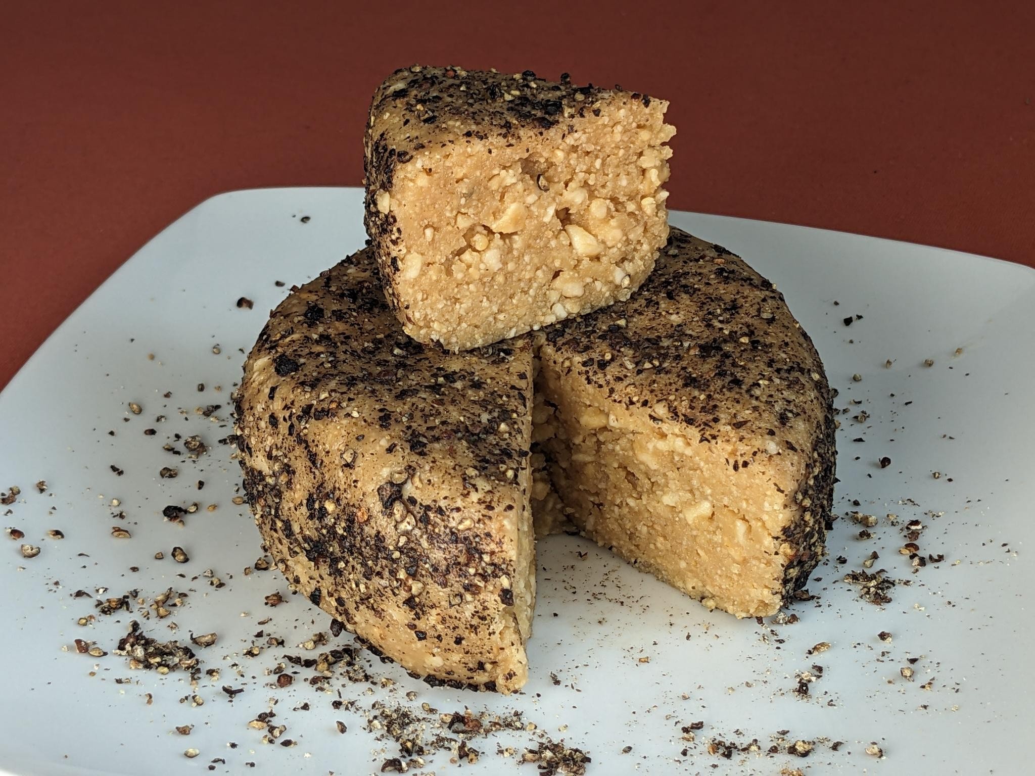 A wheel-shaped piece of cheese made from cashews on a plate. One wedge is cut out. There's ground black pepper on the outside. It looks like blinry spent a lot of time getting the lighting right for this shot.