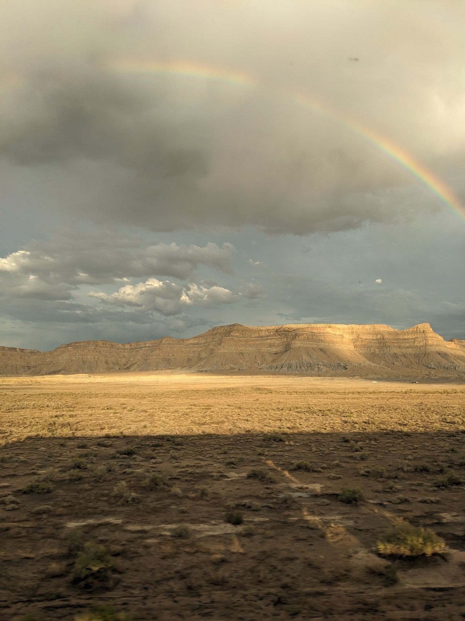 A rainbow over rocky cliffs, overseeing dry land. The shadow of the train falls onto the landscape.