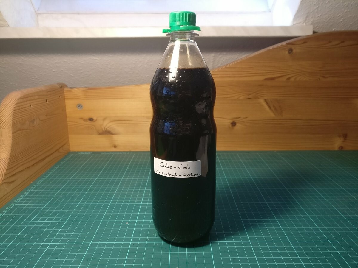 A 1 L bottle of dark brown liquid. A hand-written label says "Cube-Cola with Cyclamate + Caccharin".