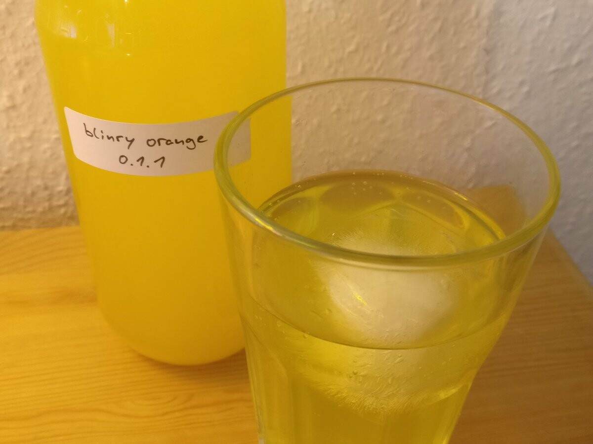 Yellow fluid in a bottle labelled "blinry orange 0.1.1". Next to it is a glass with transparent yellow fluid and an ice cube.